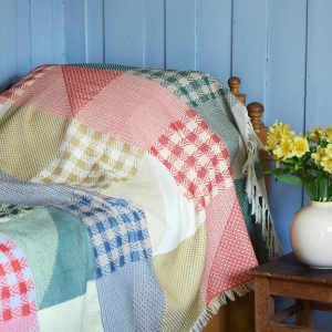 chequerd patchwork throw in nostalgic colour mix over a bed. blue walls, flowers on a stol beside the bed.