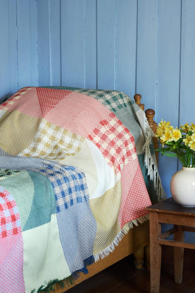 chequerd patchwork throw in nostalgic colour mix over a bed. blue walls, flowers on a stol beside the bed.