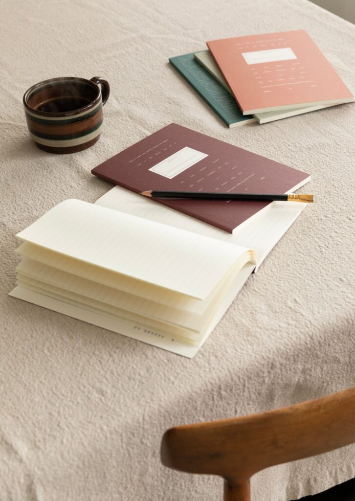 Marit Larsen x All Pine Press notebook lifestyle - colored numbered notebook editions on a table with a coffee mug and pencil